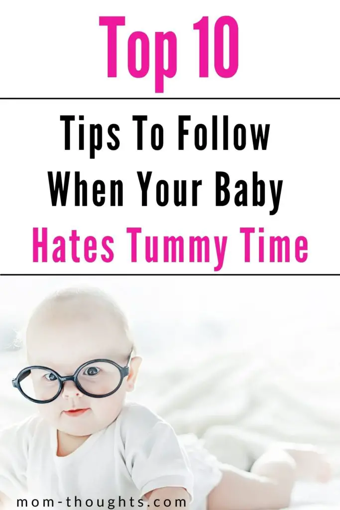 This image is of a baby with black glasses on. The baby is doing tummy time while wearing a white onesie. There is text on the top of the image that says "Top 10 tips to follow when your baby hates tummy time."