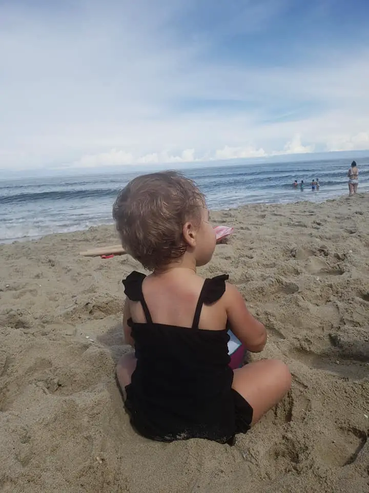 This image shows a baby girl sitting on the beach in a black baby bathing suit