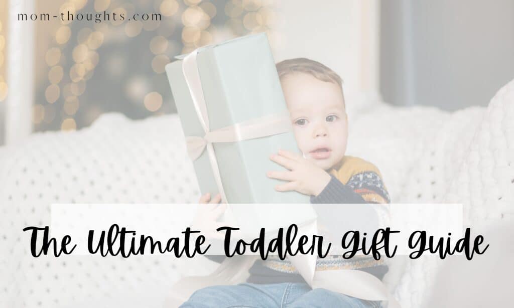 This image is of a toddler boy sitting on a couch holding a present up to his face. There is text overlay that says "The Ultimate Toddler Gift Guide". The image is on a post that lists out the best gifts for toddlers.