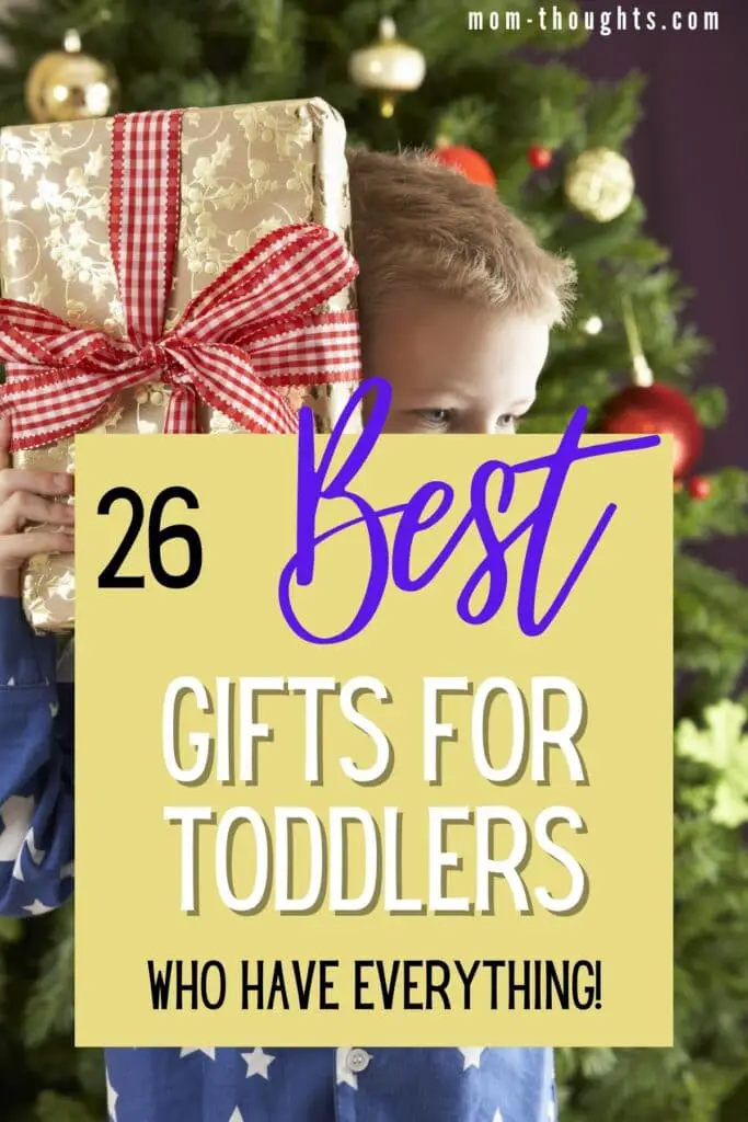 This image is of a toddler boy holding a present up to his face. The present has a big red ribbon. There is text overlay that says "26 best gifts for toddlers who have everything!"