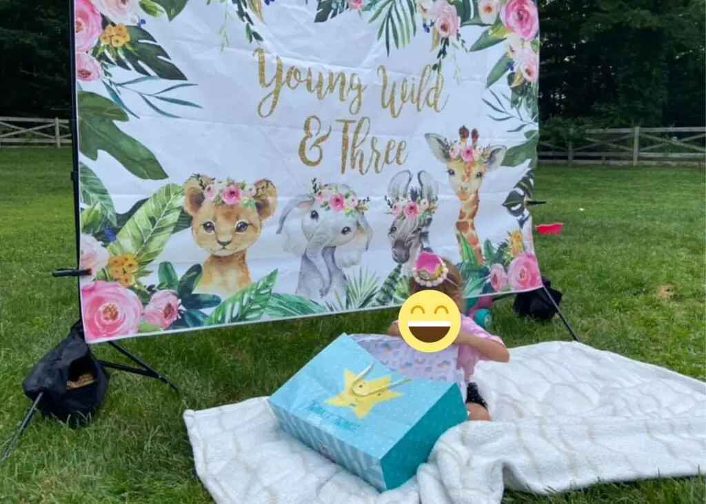 This image shows a three year old opening presents for their 3rd birthday party. They are in front of a backdrop that says "young wild and three"