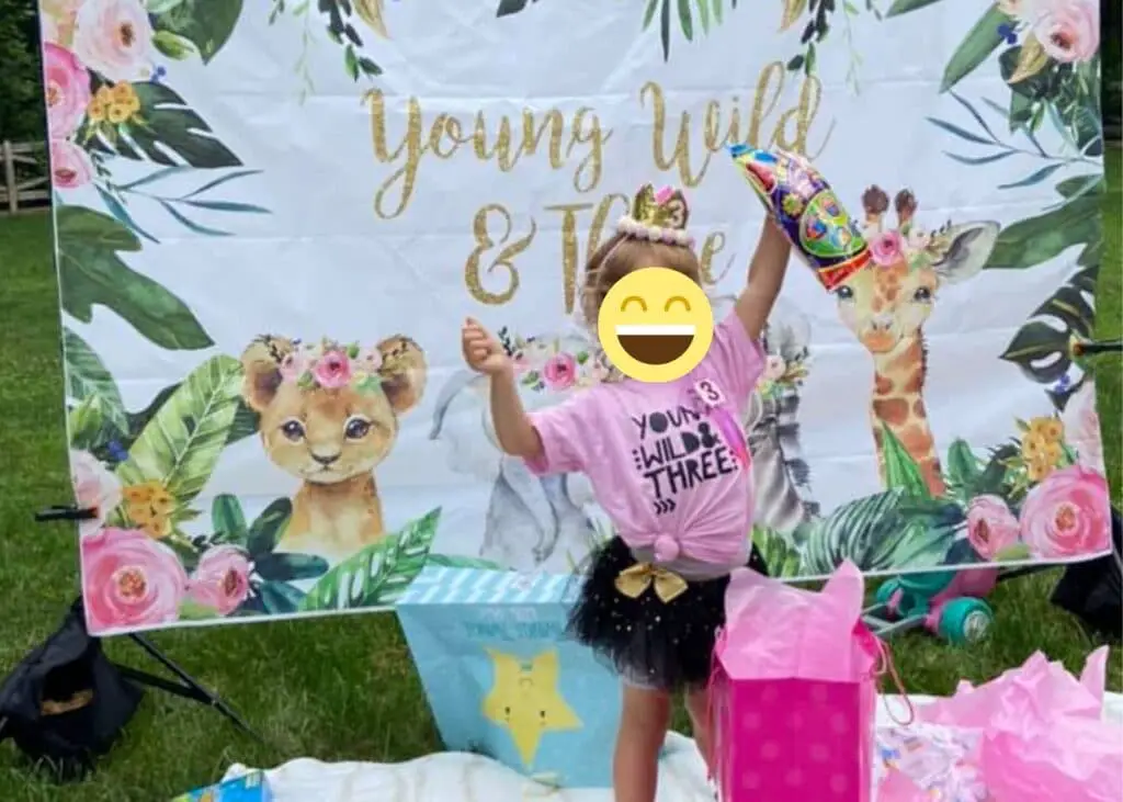 This image shows a little girl opening gifts, wearing a pink "Young Wild and Three" birthday shirt and black tutu. She's standing in front of a banner/backdrop that says Young Wild and Three. Her face is covered with a smiley face. 