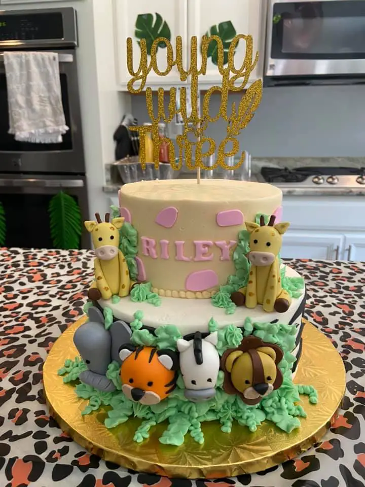 This is an image of a Young Wild and Three Birthday Cake. It's a two tier birthday cake for a third birthday party with fondant animals (elephant, tiger, zebra, lion and giraffes) sitting on the cake. The top tier has the three year old's name on it. There's a gold glitter cake topper that says "Young Wild & Three"